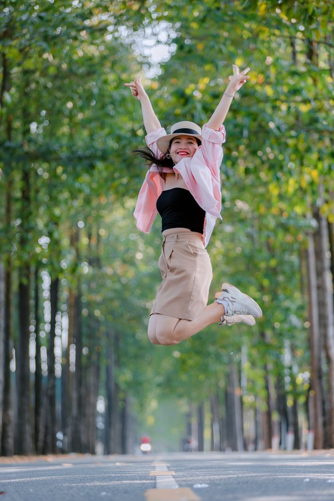 Excited young woman jumps with joy on a sunny tree-lined street, embracing outdoor happiness.